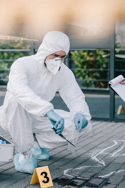 Criminologist in protective suit and latex gloves taking knife into flask at crime scene — Stock Photo