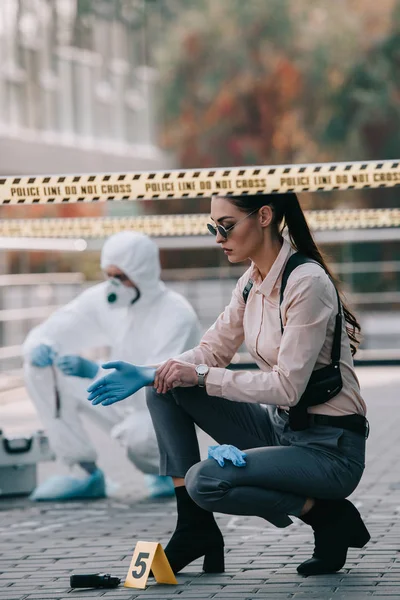 Detective mujer en gafas de sol con guantes de látex con criminólogo detrás en la escena del crimen - foto de stock