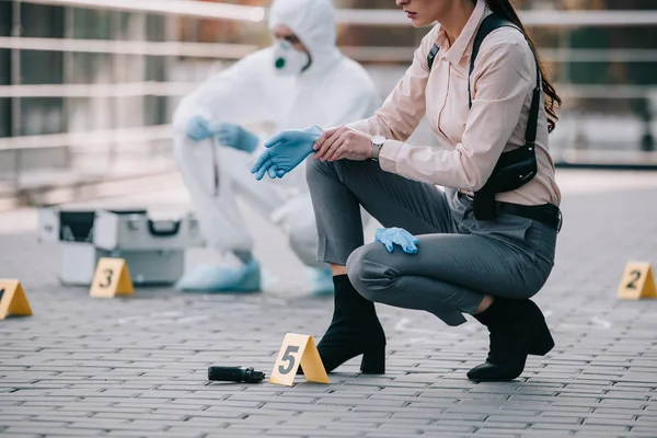 Cropped view of female detective wearing latex gloves with criminologist behind at crime scene — Stock Photo