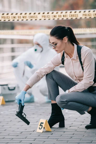 Detective donna prendere pistola come indizio con criminologo dietro di lei sulla scena del crimine — Foto stock