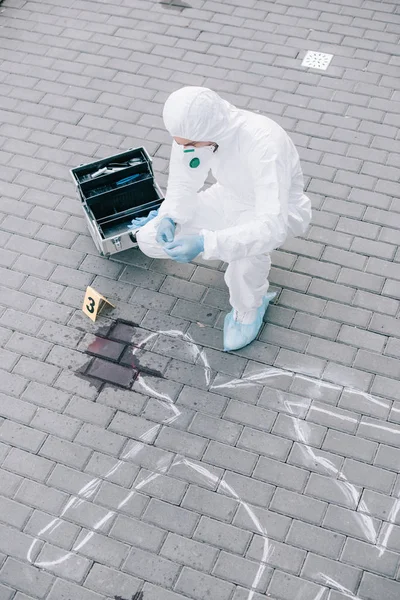 Criminólogo masculino con traje protector y guantes de látex mirando la línea de tiza con mancha de sangre en la escena del crimen - foto de stock