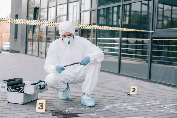 Криминалист в защитном костюме и латексных перчатках исследует орудие убийства на месте преступления — стоковое фото