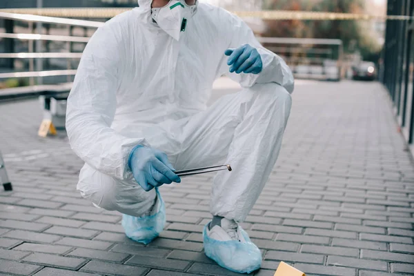 Cropped view of male criminologist in protective suit and latex gloves holding tweezers with evidence at crime scene — Stock Photo