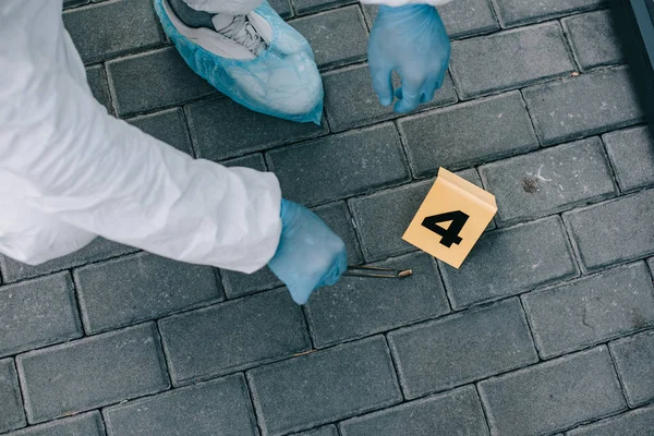 Vista parcial del criminólogo en traje de protección y guantes de látex poniendo evidencia en frasco por pinzas en la escena del crimen - foto de stock