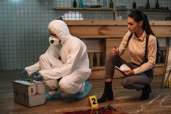 Forensic investigator and focused female detective taking notes and examining crime scene together — Stock Photo