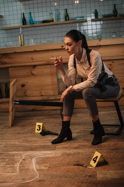 Détective féminine fumeuse concentrée assise sur la scène de crime avec des marqueurs de preuves et des contours de corps mort — Photo de stock