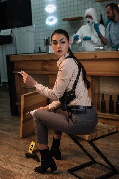 Cropped view of smoking female detective sitting at crime scene with colleagues working behind — Stock Photo