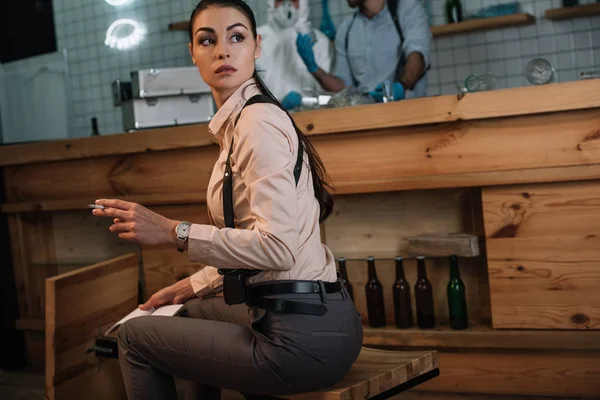 Cropped view of smoking female detective sitting at crime scene with colleagues working behind — Stock Photo