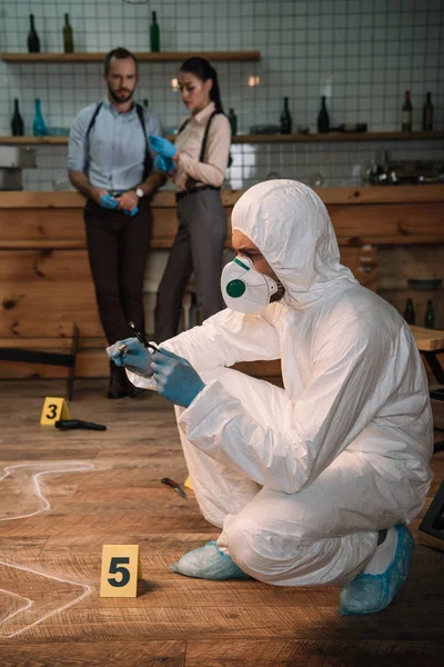 Concentarted forensic investigator examining evidence with magnifying glass at crime scene with colleagues working behind — Stock Photo