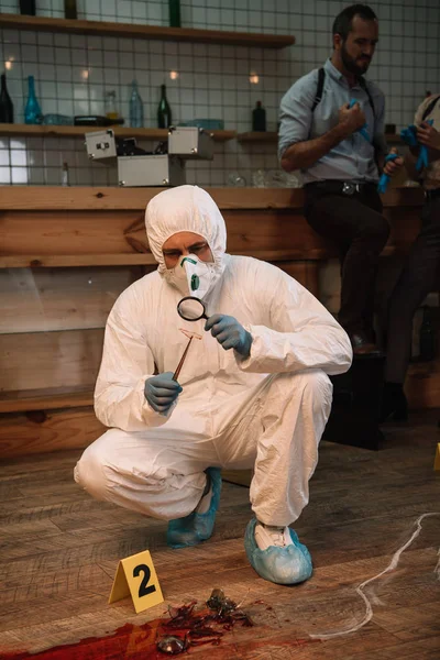 Focused forensic investigator examining evidence with magnifying glass at crime scene with colleague behind — Stock Photo