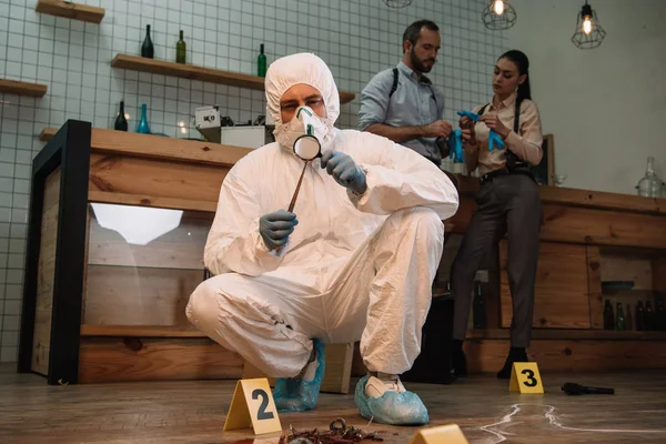 Investigador forense examinando evidencia con lupa en la escena del crimen con colegas trabajando detrás - foto de stock