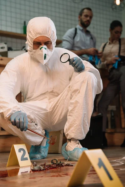 Concentarted forensic investigator examining evidence with magnifying glass at crime scene with colleagues working behind — Stock Photo