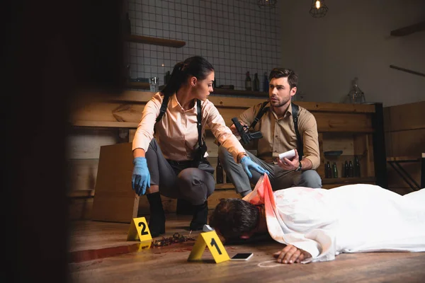 Serious female and male detectives investigating dead body at crime scene with evidence markers — Stock Photo