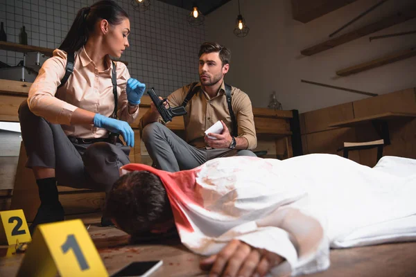 Serious female and male detectives investigating dead body at crime scene with evidence markers — Stock Photo