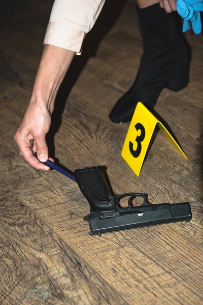 Vista recortada de la mano examinando arma cerca de marcador de evidencia en la escena del crimen - foto de stock