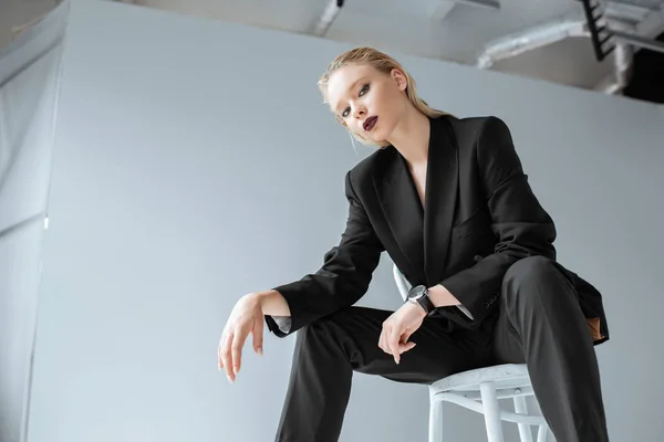 Belle jeune femme blonde en costume noir tendance assise sur chaise sur gris — Photo de stock