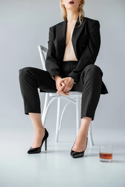 Cropped view of fashionable half naked woman in black trendy suit sitting on chair with glass of whiskey, on grey — Stock Photo