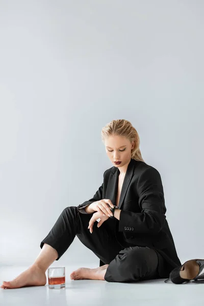 Attrayant fille en costume élégant regardant la montre tout en étant assis sur gris avec verre de cognac et talons hauts — Photo de stock