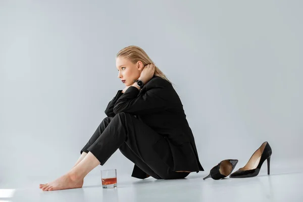 Schöne modische Mädchen in schwarzen trendigen Anzug sitzt auf grau mit einem Glas Whiskey und High Heels — Stockfoto