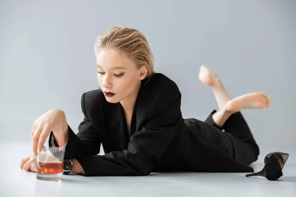 Attractive fashionable woman in black trendy suit holding glass of whiskey and lying on grey — Stock Photo