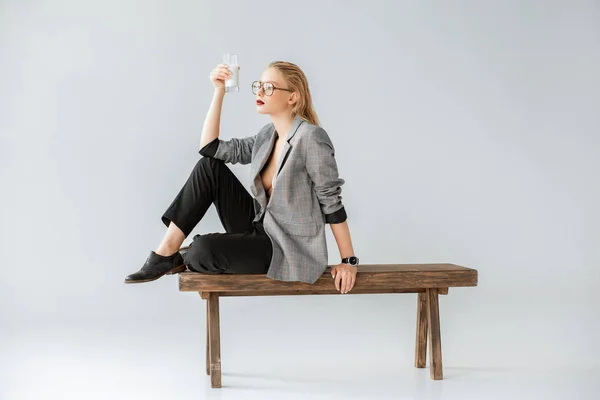 Stylish girl holding glass of milk and sitting on wooden bench on grey — Stock Photo