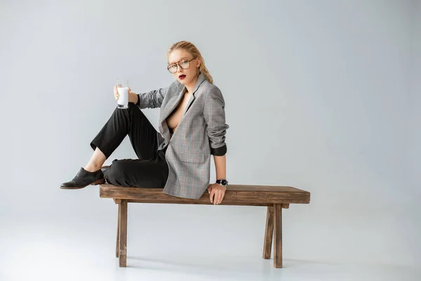 Chica de moda en chaqueta gris sosteniendo vaso de leche y sentado en banco de madera en gris - foto de stock