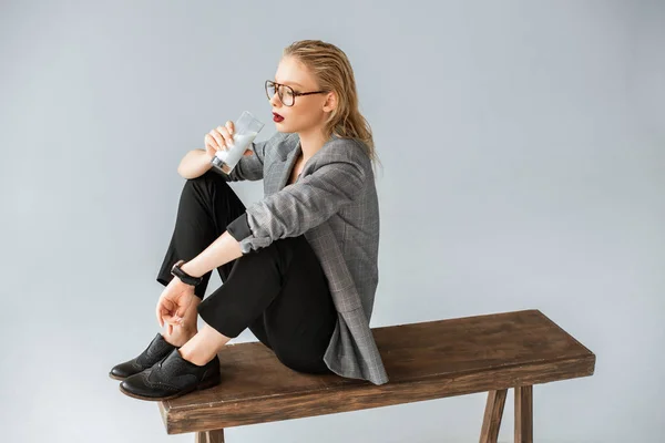 Hermosa mujer de moda beber leche de vidrio mientras está sentado en banco de madera aislado en gris - foto de stock