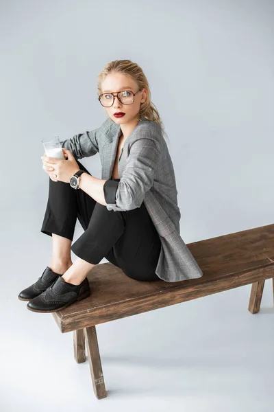 Hermosa chica en chaqueta gris sosteniendo vaso de leche y sentado en banco de madera en gris - foto de stock