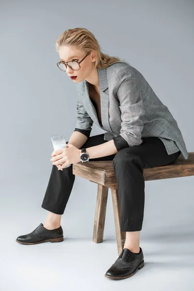 Fashionable blonde woman holding glass of milk and sitting on wooden bench on grey — Stock Photo