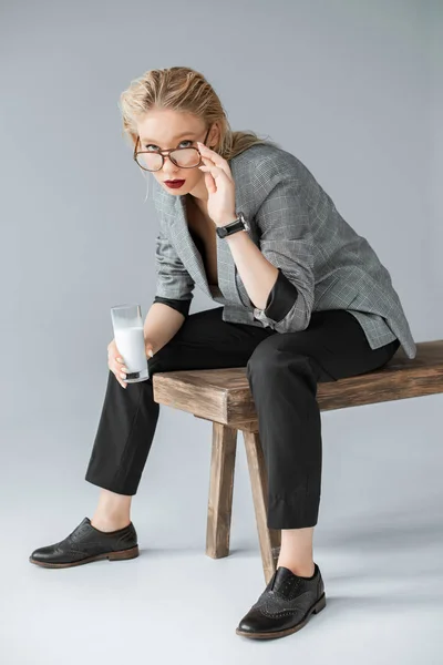 Chica de moda en gafas con vaso de leche y sentado en un banco de madera en gris - foto de stock