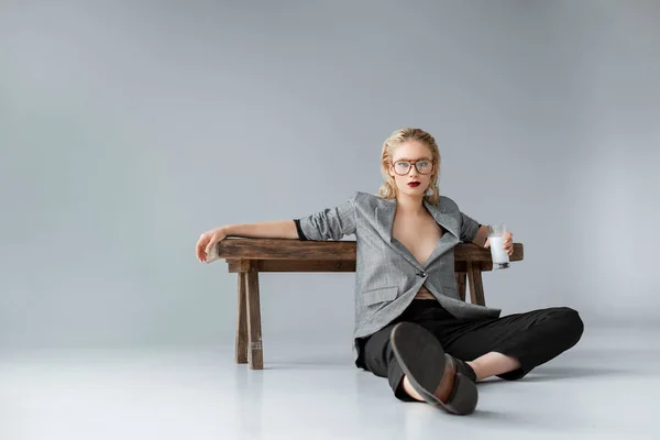 Elegante ragazza che tiene un bicchiere di latte e seduta vicino a una panchina di legno sul grigio — Foto stock