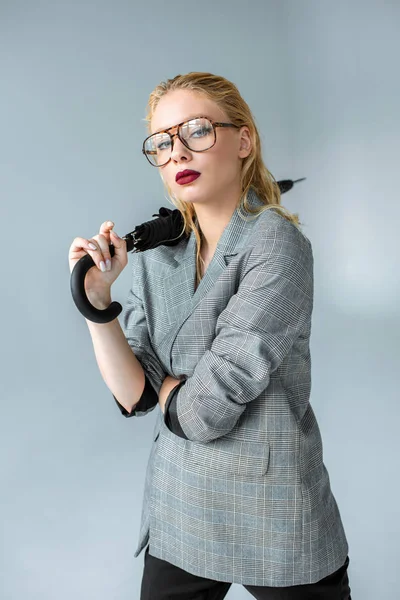 Элегантная девушка в модных очках и сером пиджаке позирует с зонтиком, изолированным на сером — стоковое фото