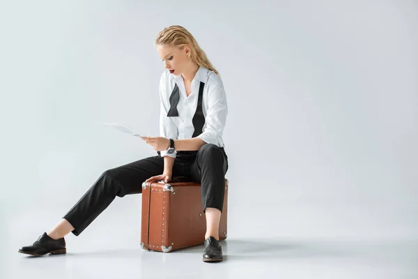 Attractive blonde girl reading newspaper while sitting on retro suitcase on grey — Stock Photo