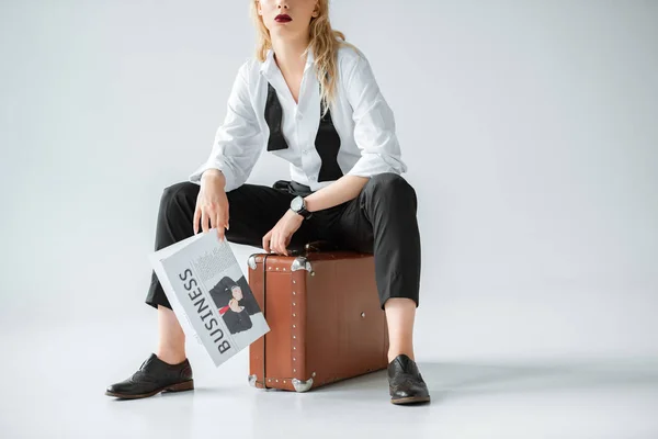 Cropped view of stylish girl holding business newspaper while sitting on retro suitcase on grey — Stock Photo