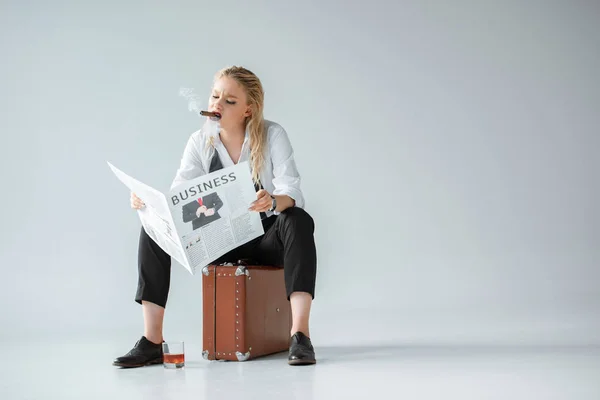 Модная девушка с сигарой и стаканом виски читает деловую газету, сидя на ретро-чемодане на сером — стоковое фото