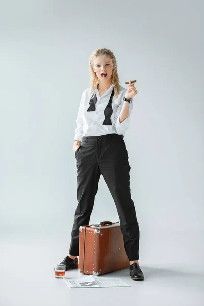 Attractive fashionable girl smoking cigar while standing on grey with vintage travel bag, newspaper and glass of whiskey — Stock Photo