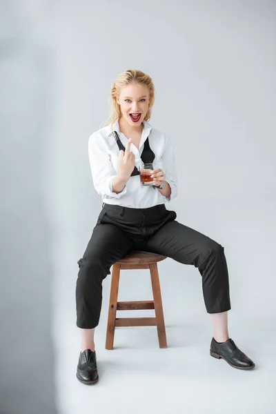Modelo de moda excitado con vaso de whisky mostrando el dedo medio mientras está sentado en el taburete en gris - foto de stock