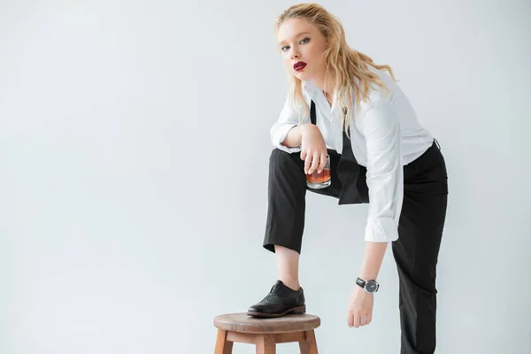 Attraktives blondes Model mit Glas Whiskey posiert mit Hocker, isoliert auf grau — Stockfoto