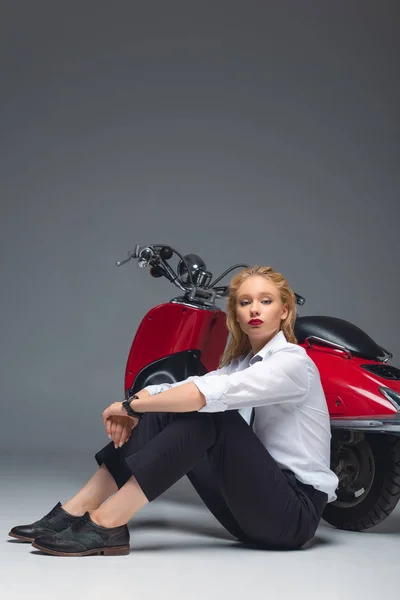 Elegante chica de moda sentado cerca de scooter rojo en gris - foto de stock