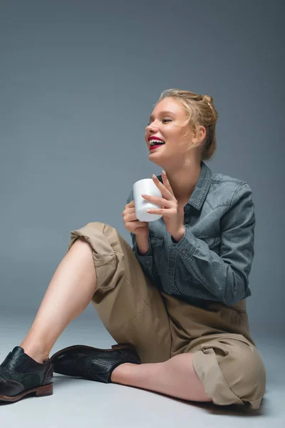 Attractive stylish laughing girl with cup of tea sitting on grey — Stock Photo