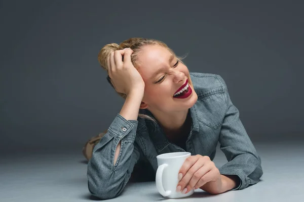 Schönes lachendes Mädchen mit einer Tasse Kaffee auf grau liegend — Stockfoto