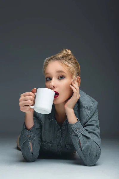 Attractive surprised girl drinking tea from white cup on grey — Stock Photo