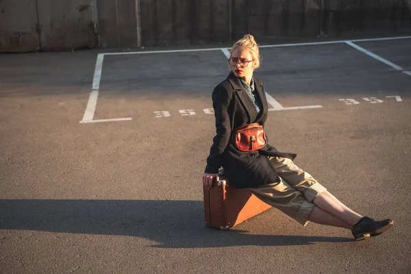 Fashionable beautiful girl sitting on vintage travel bag on urban parking — Stock Photo