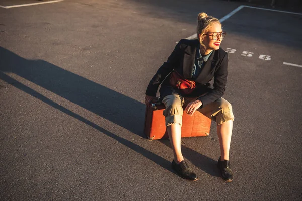 Модна усміхнена дівчина сидить на ретро валізі на міській парковці — стокове фото