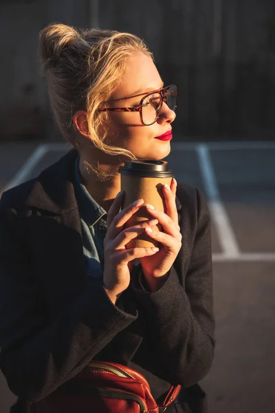 Dreamy blonde woman holding disposable cup of coffee on parking — Stock Photo