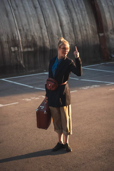 Ragazza sorridente alla moda con valigia vintage che mostra il dito medio sul parcheggio urbano — Foto stock