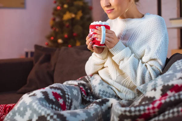 Vue recadrée d'une jeune femme couverte d'une couverture assise sur un canapé et tenant une tasse de cacao chaud avec des guimauves à Noël — Photo de stock