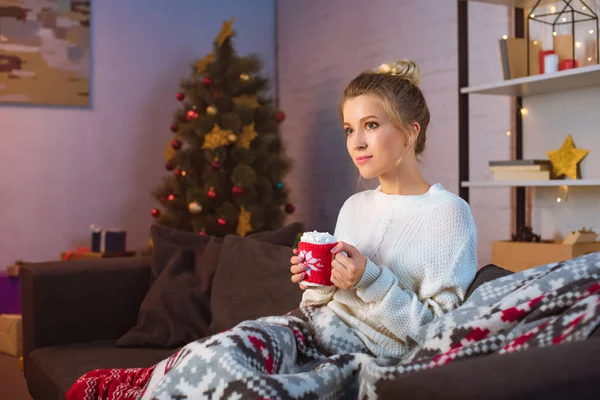 Mujer rubia joven cubierta de manta sentada en el sofá y sosteniendo la taza de cacao caliente con malvaviscos en Navidad — Stock Photo