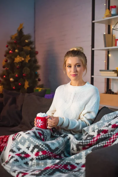 Young blonde woman sitting on couch, holding cup of hot cocoa with marshmallows and looking at camera at christmas time — Stock Photo
