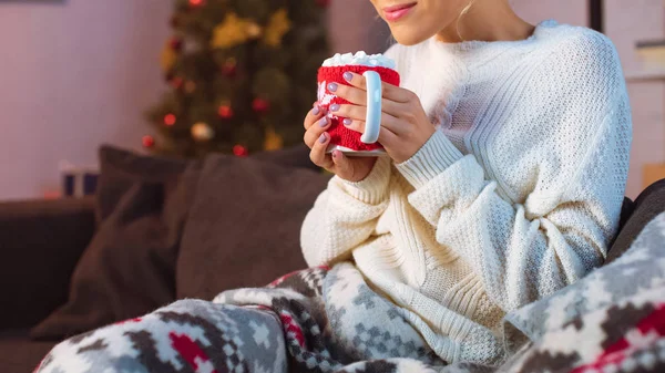 Vue recadrée d'une jeune femme couverte d'une couverture assise sur un canapé et tenant une tasse de cacao chaud avec des guimauves à Noël — Photo de stock